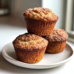 Three golden brown sourdough discard banana muffins stacked and resting on a white plate, topped with cinnamon sugar.