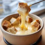 A piece of crusty bread is lifted from a bowl of velvety cheese fondue, showing a long, satisfying cheese pull.