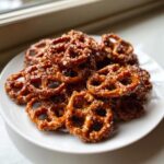 A close-up of a white plate piled high with shiny, glazed toffee pretzels sprinkled with coarse sea salt.