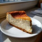 A thick slice of creamy white chocolate cheesecake with a golden-brown top, served on a white plate.