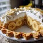 A slice removed from an amazing banana cream pie showing layers of crust, custard, bananas, and whipped cream topping.