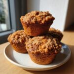 A stack of four golden brown apple cinnamon muffins topped with a generous streusel crumb topping, sitting on a plate.