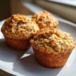 Three golden brown banana oatmeal muffins sitting on a light gray plate, highlighted by natural sunlight.