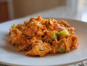 A close-up serving of vibrant orange buffalo chicken salad mixed with celery and red onion on a white plate.