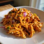 A close-up of vibrant, shredded buffalo chicken salad mixed with red onion and green peppers served on a white dish.