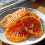 Close-up of bright orange, glistening candied orange slices coated in sugar on a white plate.