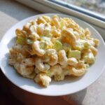 A close-up of creamy amish macaroni salad with elbow pasta and chopped celery served on a white plate.