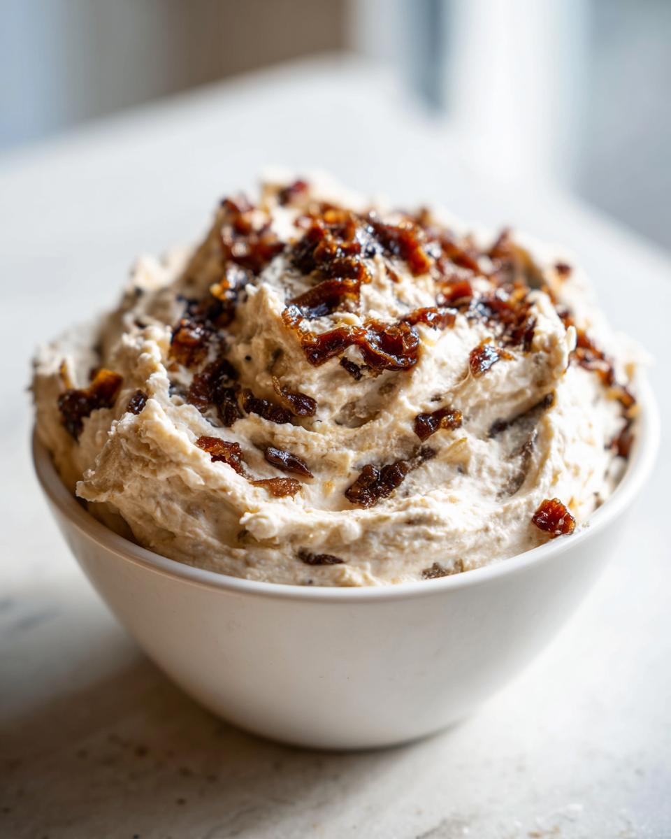 A close-up of creamy caramelized onion dip mounded high in a white bowl, topped with dark, rich caramelized onions.