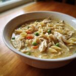 A close-up of a hearty bowl of creamy chicken noodle soup filled with shredded chicken, egg noodles, peas, and carrots.