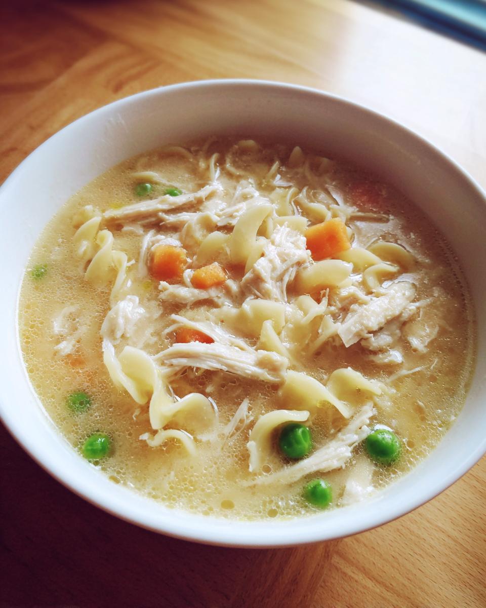 Close-up of a white bowl filled with creamy chicken noodle soup, featuring shredded chicken, egg noodles, peas, and carrots.
