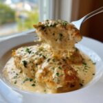 A fork lifts a piece of chicken covered in rich, creamy garlic chicken sauce, garnished with parsley.