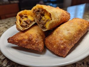 Four crispy, golden-brown cheeseburger egg rolls stacked on a white plate, one cut open showing seasoned ground beef and melted cheddar cheese.
