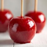 A close-up of a perfectly glossy, bright red candy apple on a wooden stick, with two more in the background.