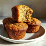 A stack of four golden-brown protein muffins on a white plate, with the top one broken open to show the moist interior.