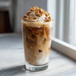 A tall glass of iced cookie butter latte with swirling milk and espresso, topped with whipped cream and cookie crumbles.