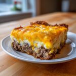 A thick slice of meatloaf casserole topped with mashed potatoes and melted cheddar cheese on a white plate.