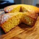 Close-up of a slice of moist, golden cornbread showing its crumb texture, with the rest of the skillet cornbread in the background.