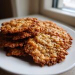 A stack of perfectly baked, golden brown oatmeal lace cookies resting on a white plate near a window.