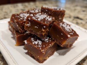 Close-up of rich, layered salted caramel fudge squares topped with flaky sea salt on a white plate.