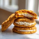 A stack of three soft pumpkin cheesecake cookies filled with cream cheese frosting, one cookie is cut in half on top.