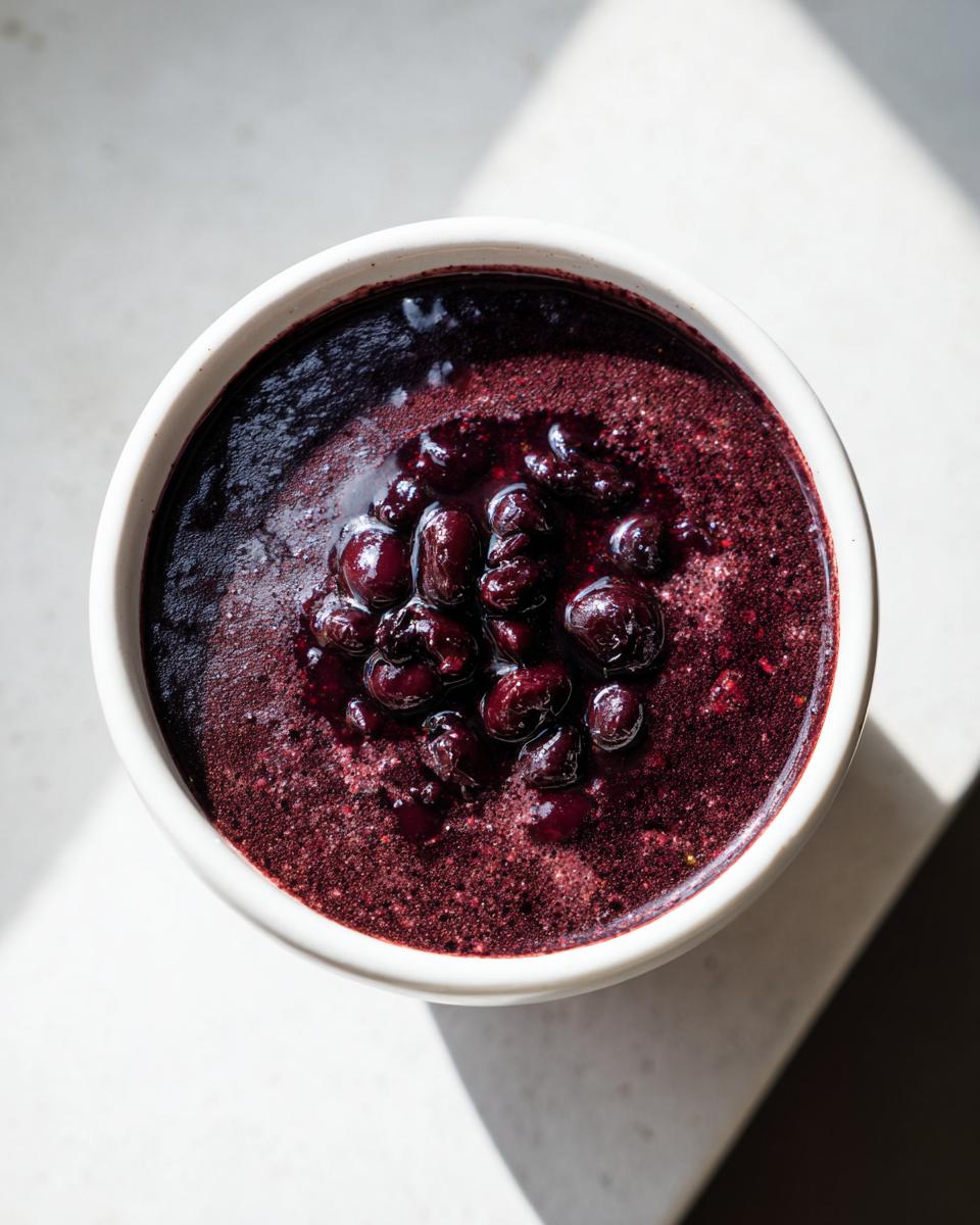 A bowl of dark purple, thick puree topped with whole, glossy berries, contrasting with the bright white bowl and background.