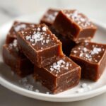 Close-up of rich, brown salted caramel fudge squares topped with flaky sea salt on a white plate.