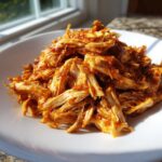 Close-up of shredded chicken tinga coated in a vibrant, rich red, smoky sauce piled on a white plate.