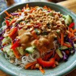 Close-up of a vibrant spring roll salad featuring rice noodles, shredded carrots, cucumber, red pepper, and a generous drizzle of peanut sauce topped with crushed peanuts.