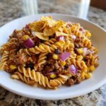 A close-up view of a serving of taco pasta salad featuring rotini pasta, seasoned ground beef, corn, black beans, and red onion, topped with crushed tortilla chips.