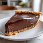 A rich, dark slice of chocolate pie with a thick, smooth filling and a crumbly graham cracker crust on a white plate.