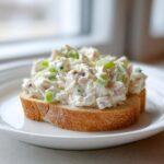 A thick scoop of creamy ham salad with visible celery chunks served open-faced on a slice of toasted bread.