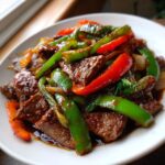 A close-up of glossy, saucy pepper steak mixed with vibrant green and red bell pepper strips on a white plate.