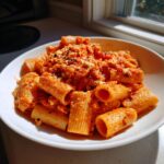 A white bowl filled with rigatoni pasta coated in a creamy, orange-red sauce and topped with crumbled feta cheese, ready for the baked feta pasta recipe.
