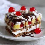 A decadent slice of banana split cake featuring layers of graham cracker crust, cream, pineapple, cherries, and topped with whipped cream and chocolate syrup.