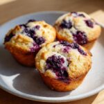Three golden-brown blueberry cottage cheese muffins with burst blueberries on a speckled white plate.