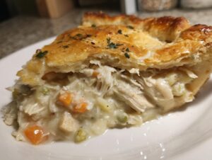 Close-up of a slice of chicken pot pie showing shredded chicken, carrots, and peas in a creamy sauce under a golden, flaky crust.