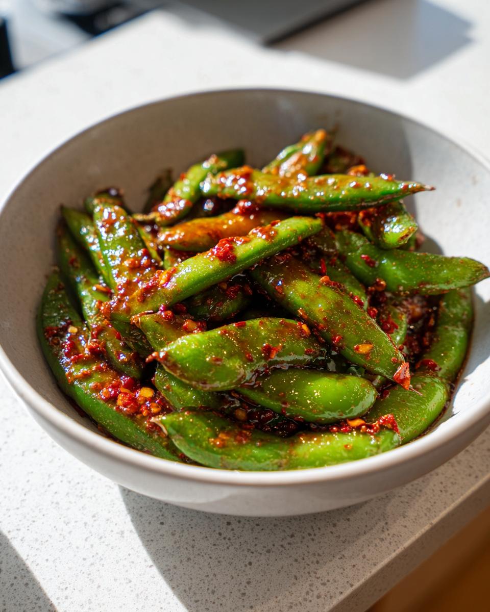 Close-up of bright green snap peas coated in a glossy, spicy chili crisp dressing, served in a light gray bowl.