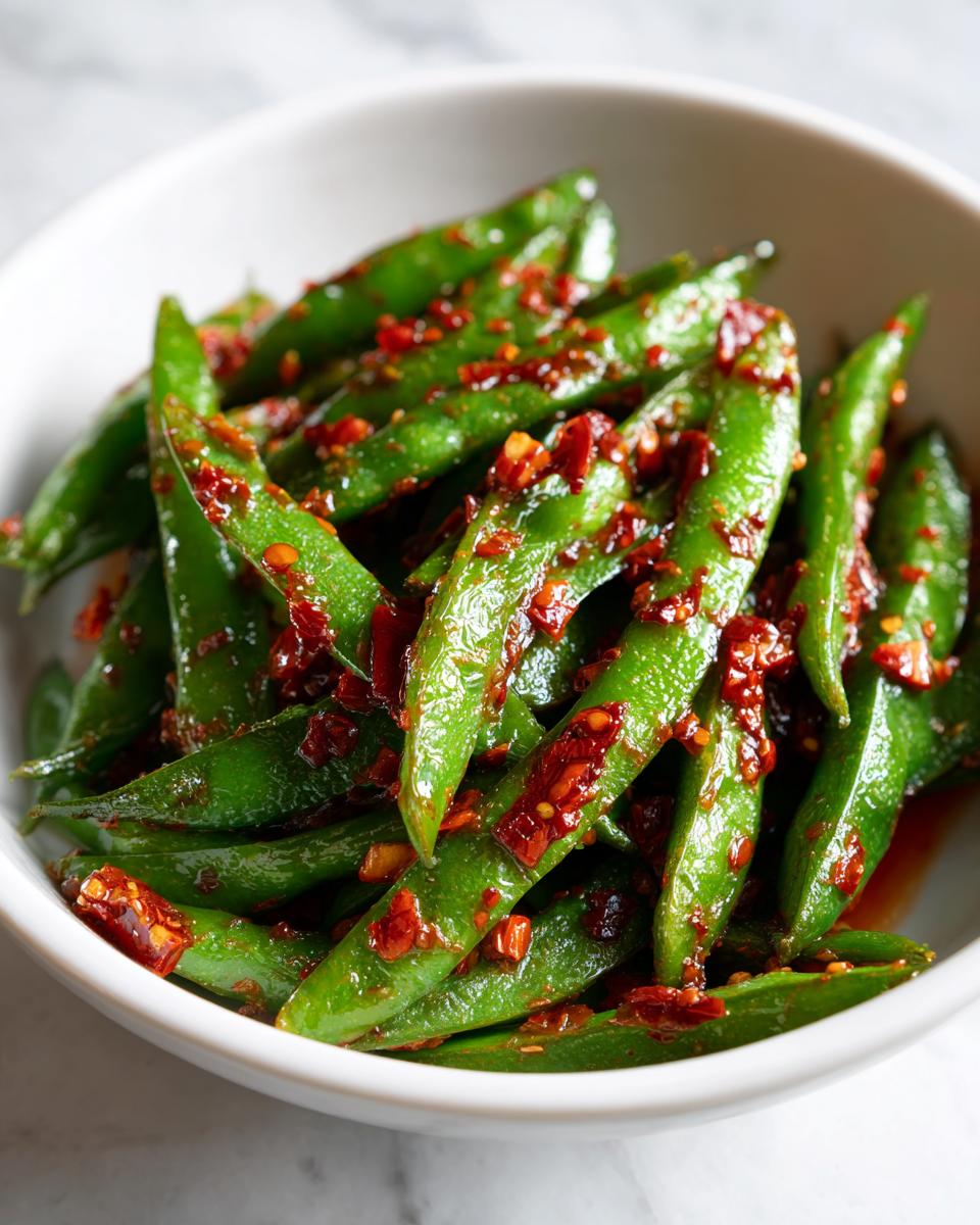 Close-up of bright green snap peas tossed generously with red chili crisp, served in a white bowl.