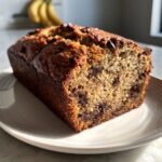 A golden brown loaf of chocolate chip peanut butter banana bread sitting on a white plate.