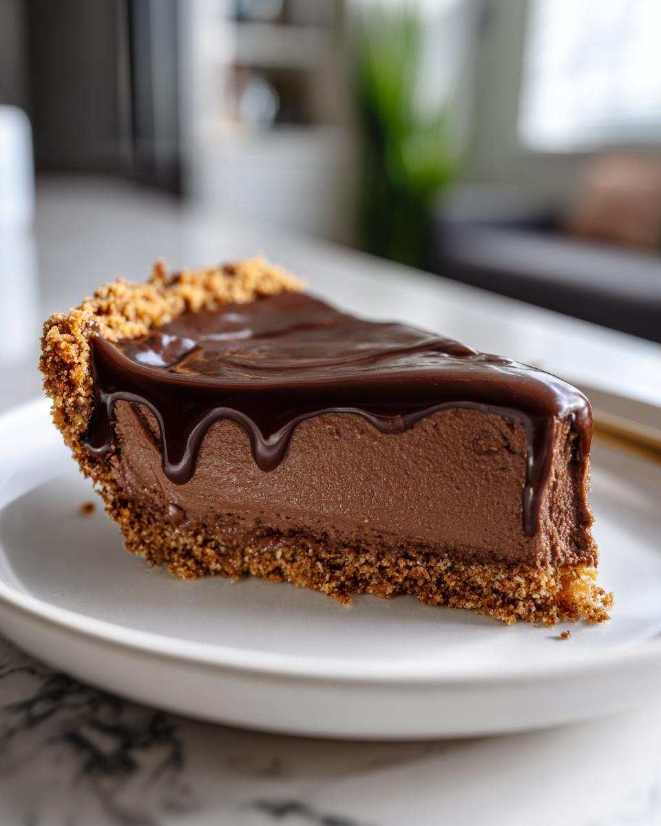 A decadent slice of chocolate pie featuring a thick, creamy filling, graham cracker crust, and glossy chocolate ganache topping.