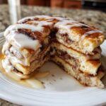 A close-up of a stack of cinnamon roll pancakes cut in half, generously topped with white icing.