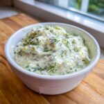 A close-up of thick, creamy dill dip generously topped with fresh dill sprigs, served in a white bowl.
