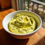 A close-up of bright green creamy jalapeño hummus swirled in a white bowl, drizzled with olive oil and topped with pepper.