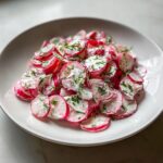 A white bowl filled with thinly sliced radish salad coated in a creamy dressing and topped with fresh chopped chives.