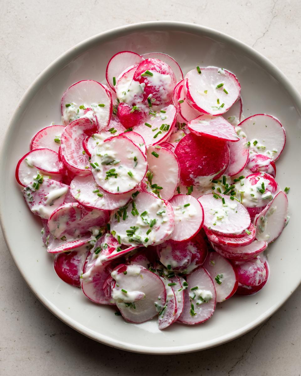 Overhead view of a creamy radish salad featuring thinly sliced red radishes coated in a white dressing and topped with fresh green chives.