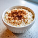 Close-up of a small white bowl filled with creamy rice pudding, generously dusted with ground cinnamon.