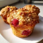 Three golden brown raspberry muffins with a crumb topping are displayed on a white plate.