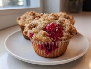 A close-up of a freshly baked raspberry muffin with a generous brown sugar crumb topping, sitting on a white plate.