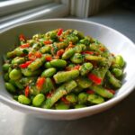 Close-up of a vibrant cucumber edamame salad mixed with red peppers and sesame seeds in a white bowl.