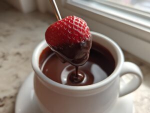 A fresh strawberry being dipped into rich, melted chocolate fondue served in a white mug.